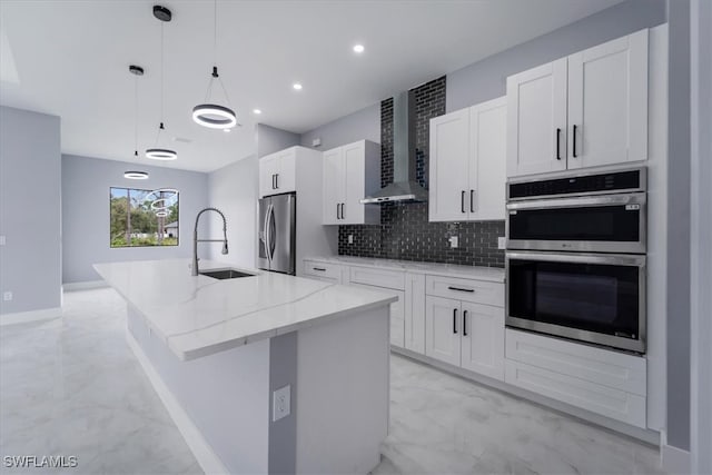 kitchen featuring wall chimney exhaust hood, stainless steel appliances, decorative light fixtures, a kitchen island with sink, and white cabinets