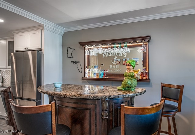 bar featuring white cabinets, decorative backsplash, dark stone countertops, ornamental molding, and stainless steel refrigerator