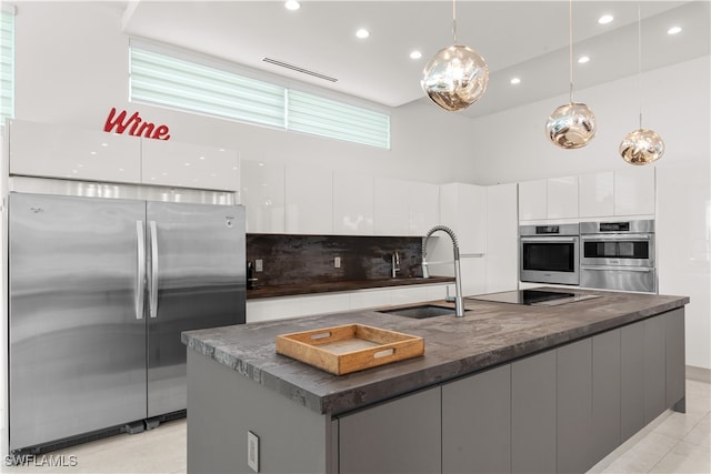 kitchen with a towering ceiling, stainless steel appliances, a kitchen island with sink, pendant lighting, and white cabinets