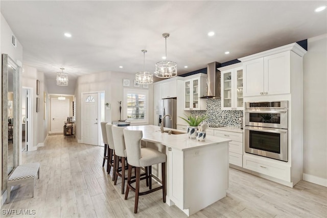 kitchen with decorative light fixtures, a center island with sink, light countertops, glass insert cabinets, and wall chimney range hood