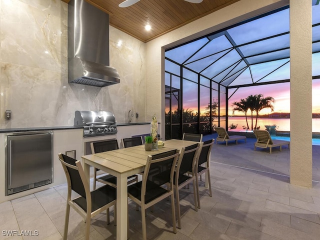 patio terrace at dusk featuring a lanai, a grill, a water view, and exterior kitchen