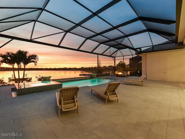 pool at dusk featuring glass enclosure, a patio area, a water view, and an in ground hot tub