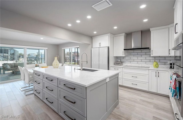 kitchen featuring a kitchen island with sink, wall chimney range hood, light stone counters, white cabinetry, and fridge