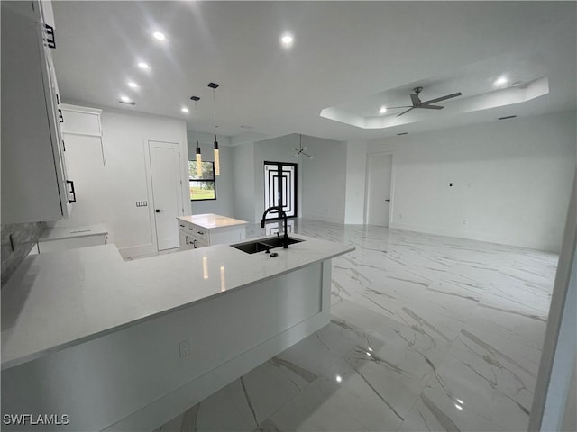 kitchen with ceiling fan, sink, a raised ceiling, a kitchen island with sink, and white cabinets