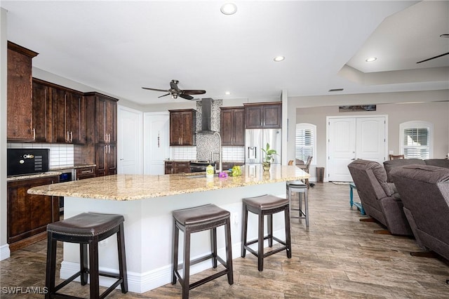 kitchen featuring ceiling fan, wall chimney range hood, tasteful backsplash, stainless steel fridge with ice dispenser, and a large island with sink