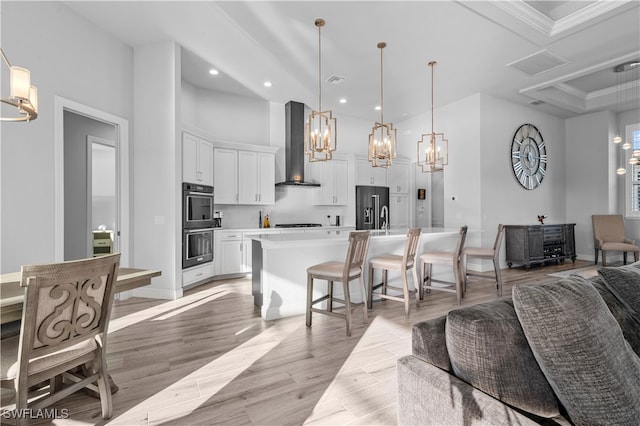 kitchen with wall chimney exhaust hood, light wood-type flooring, an island with sink, decorative light fixtures, and white cabinetry