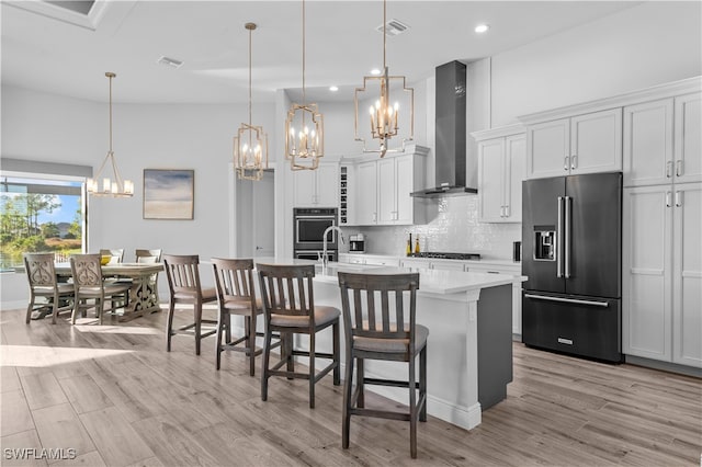 kitchen with wall chimney exhaust hood, an island with sink, decorative light fixtures, white cabinets, and appliances with stainless steel finishes