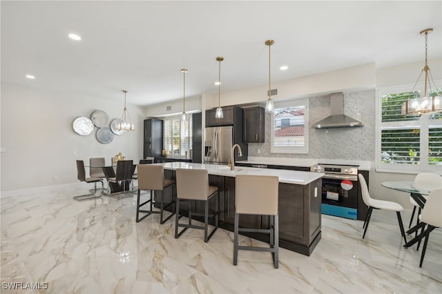 kitchen featuring pendant lighting, backsplash, a kitchen island with sink, wall chimney range hood, and appliances with stainless steel finishes