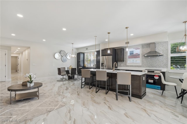 kitchen with pendant lighting, wall chimney range hood, a kitchen island with sink, and appliances with stainless steel finishes