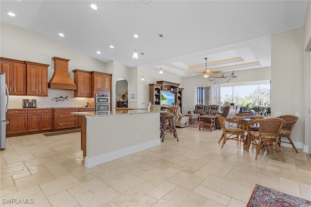 kitchen featuring custom exhaust hood, a center island with sink, ceiling fan, appliances with stainless steel finishes, and light stone counters