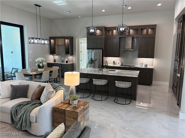 kitchen with hanging light fixtures, appliances with stainless steel finishes, light stone countertops, a kitchen island with sink, and wall chimney range hood