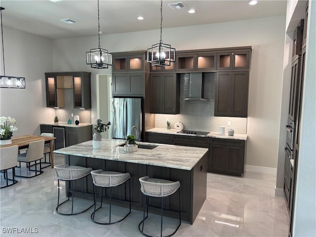 kitchen with pendant lighting, stainless steel refrigerator, a kitchen island with sink, light stone counters, and wall chimney exhaust hood