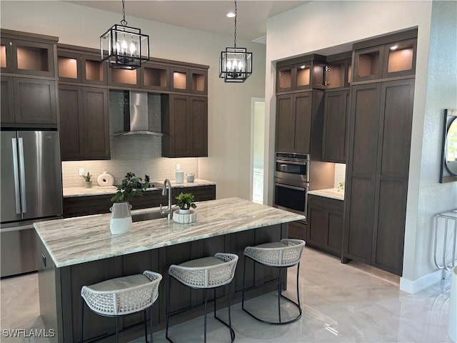 kitchen with wall chimney range hood, stainless steel appliances, light stone counters, an island with sink, and decorative backsplash