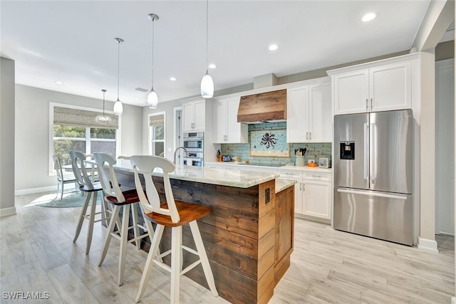 kitchen featuring premium range hood, appliances with stainless steel finishes, decorative light fixtures, white cabinetry, and a center island with sink