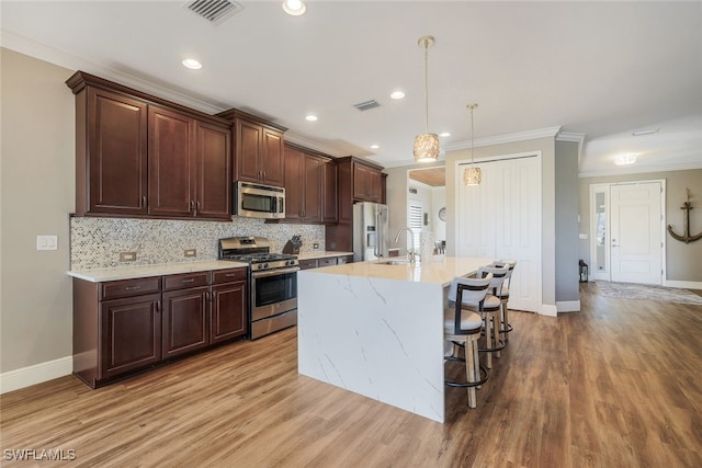 kitchen with hanging light fixtures, stainless steel appliances, crown molding, an island with sink, and light wood-type flooring