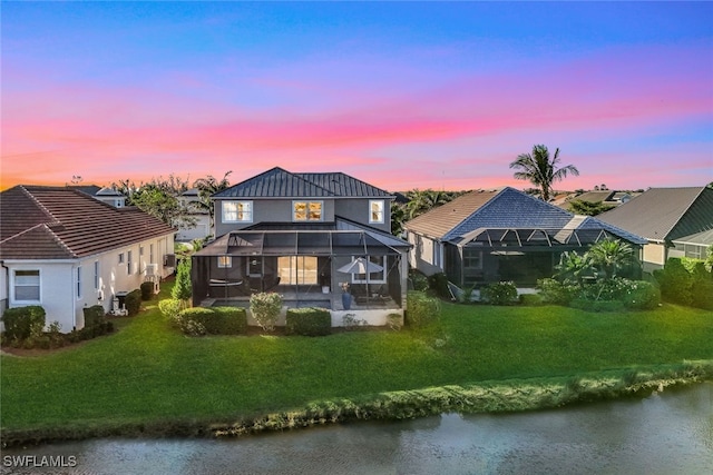 back house at dusk featuring glass enclosure, a water view, and a yard