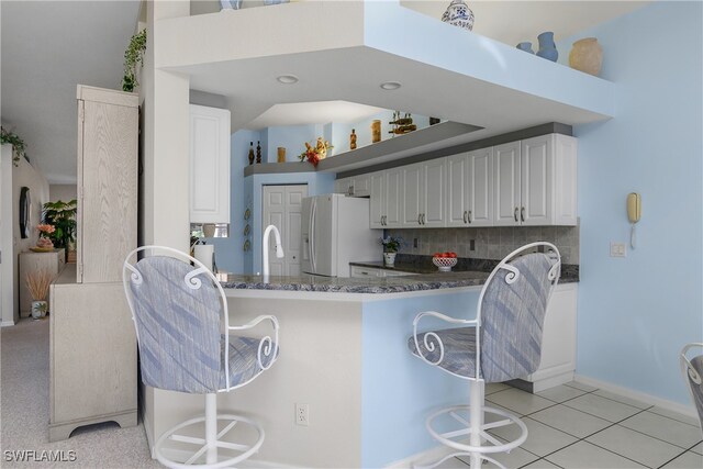 kitchen with white cabinetry, white fridge with ice dispenser, kitchen peninsula, dark stone countertops, and a breakfast bar