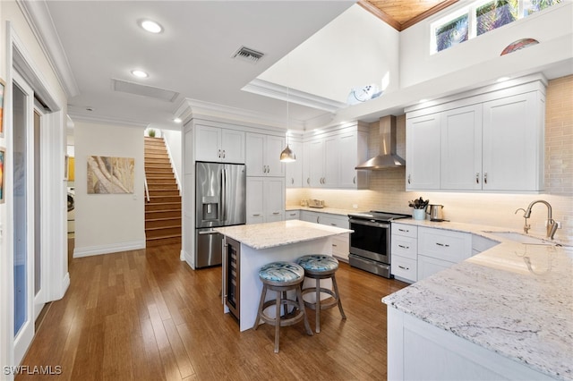 kitchen featuring wall chimney range hood, decorative light fixtures, a kitchen island, white cabinetry, and stainless steel appliances