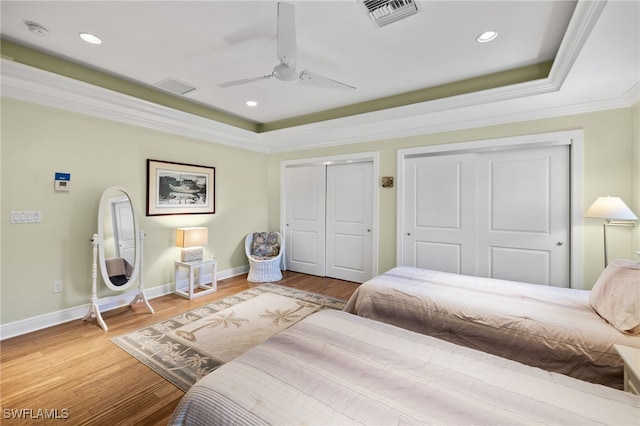 bedroom with hardwood / wood-style floors, a raised ceiling, crown molding, ceiling fan, and multiple closets