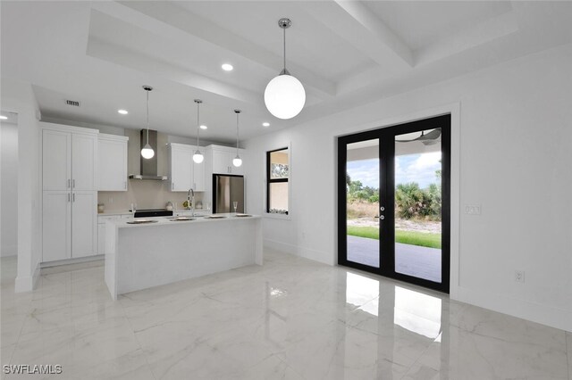 kitchen with white cabinetry, wall chimney exhaust hood, stainless steel fridge, pendant lighting, and a center island with sink