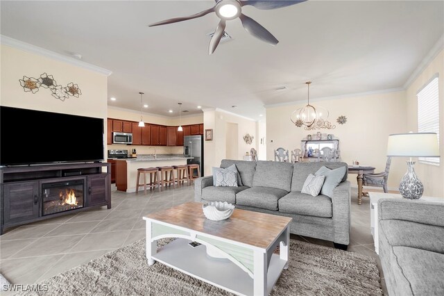 living room with light tile patterned floors, ornamental molding, ceiling fan with notable chandelier, and a glass covered fireplace