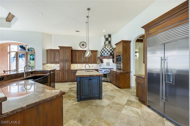 kitchen featuring high end appliances, a center island with sink, sink, hanging light fixtures, and custom range hood