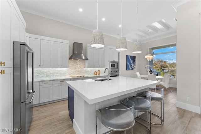 kitchen with a sink, light countertops, appliances with stainless steel finishes, wall chimney exhaust hood, and a center island with sink