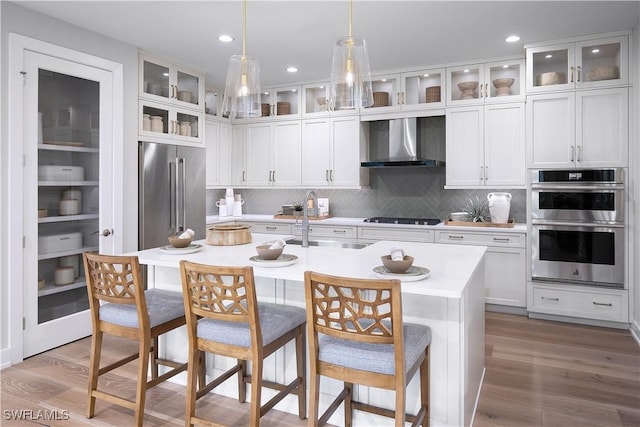 kitchen with a sink, wall chimney range hood, wood finished floors, stainless steel appliances, and a kitchen island with sink