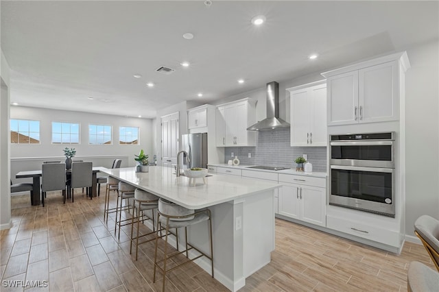 kitchen with wall chimney exhaust hood, stainless steel appliances, a center island with sink, white cabinets, and light wood-type flooring