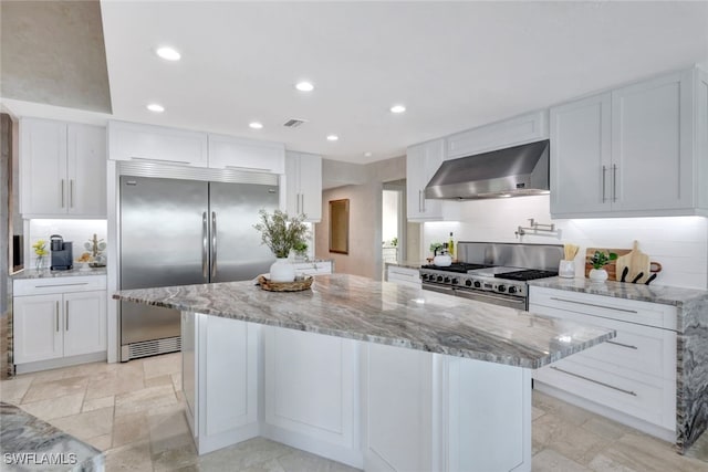 kitchen with light stone countertops, white cabinetry, high quality appliances, and wall chimney range hood