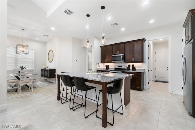 kitchen featuring light stone countertops, appliances with stainless steel finishes, dark brown cabinetry, a center island with sink, and decorative light fixtures