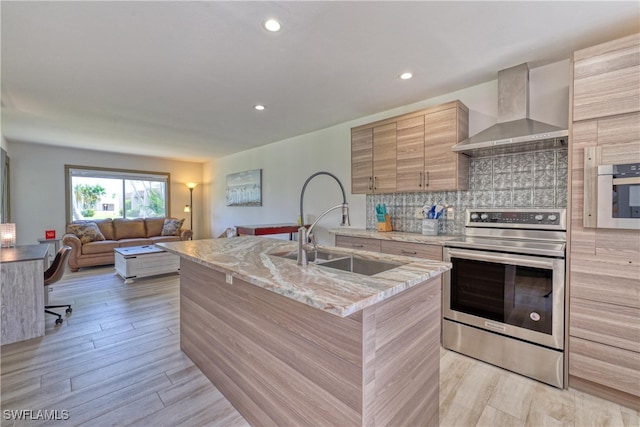 kitchen featuring wall chimney exhaust hood, stainless steel appliances, light hardwood / wood-style flooring, backsplash, and an island with sink