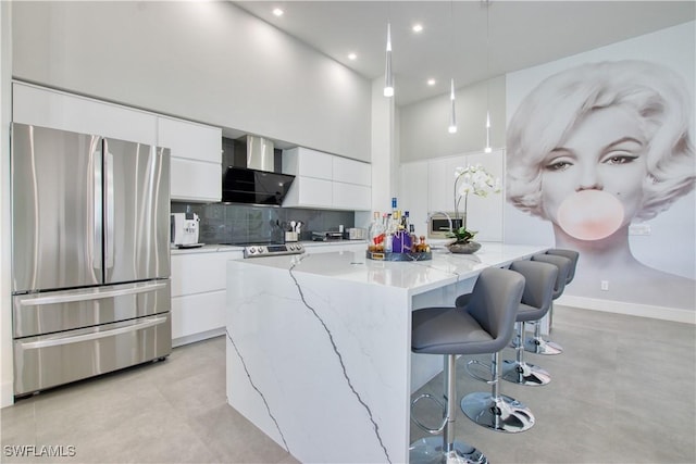 kitchen featuring wall chimney exhaust hood, pendant lighting, a kitchen island with sink, white cabinets, and appliances with stainless steel finishes