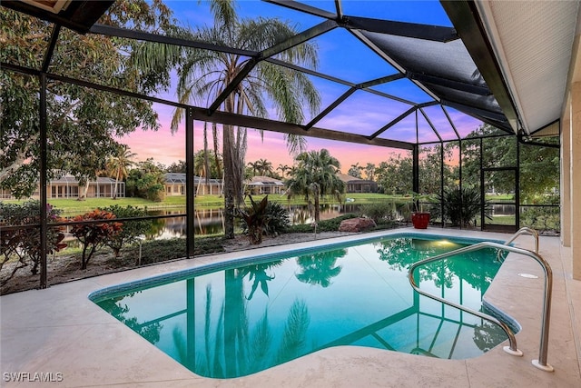 pool at dusk with glass enclosure, a patio area, and a water view