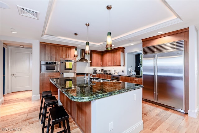 kitchen with hanging light fixtures, stainless steel appliances, an island with sink, wall chimney exhaust hood, and a tray ceiling