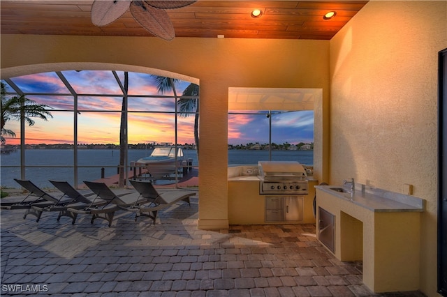 patio terrace at dusk with glass enclosure, a water view, ceiling fan, area for grilling, and exterior kitchen