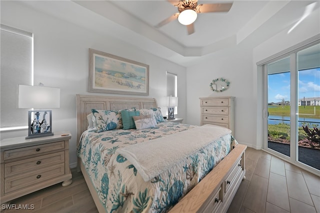 bedroom featuring access to exterior, a tray ceiling, ceiling fan, dark wood-type flooring, and a water view