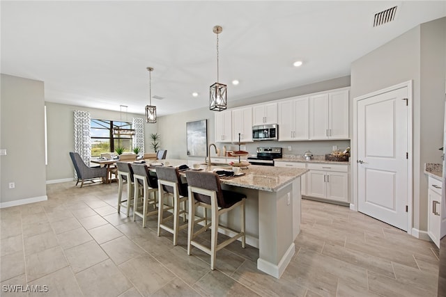 kitchen with light stone counters, decorative light fixtures, a center island with sink, white cabinets, and appliances with stainless steel finishes