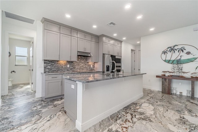 kitchen featuring backsplash, gray cabinetry, sink, a center island with sink, and dark stone countertops