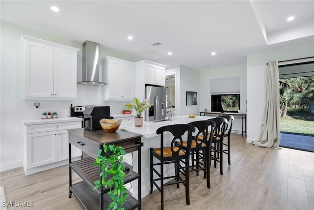 kitchen with white cabinets, appliances with stainless steel finishes, light wood-type flooring, wall chimney exhaust hood, and a center island with sink