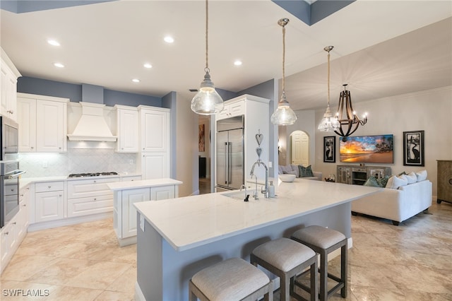 kitchen featuring custom exhaust hood, sink, pendant lighting, built in appliances, and an island with sink