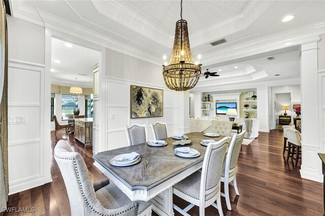 dining space featuring dark wood-type flooring, ceiling fan with notable chandelier, built in features, ornamental molding, and a tray ceiling