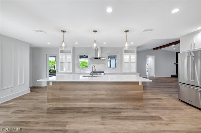 kitchen featuring pendant lighting, high end refrigerator, wall chimney range hood, an island with sink, and light hardwood / wood-style floors