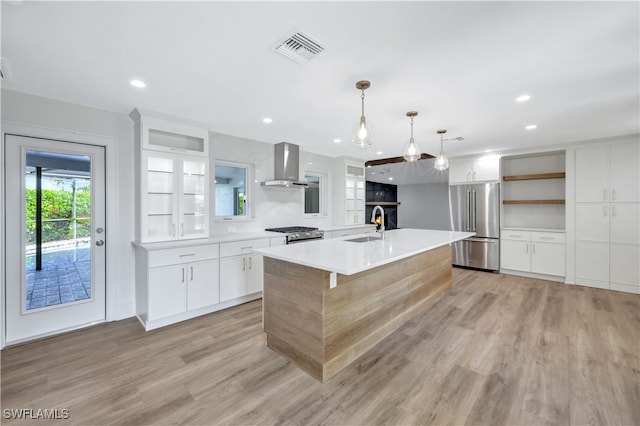 kitchen featuring pendant lighting, a center island with sink, wall chimney range hood, appliances with stainless steel finishes, and white cabinetry