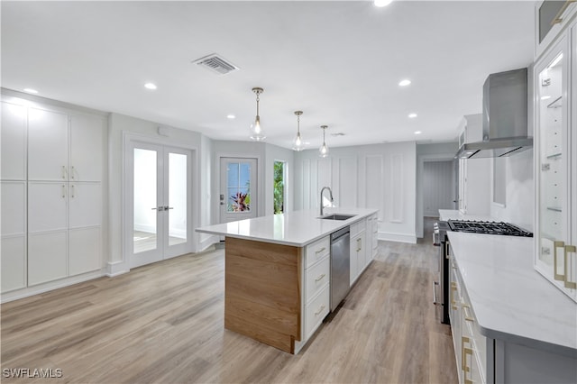 kitchen with wall chimney exhaust hood, hanging light fixtures, stainless steel appliances, a kitchen island with sink, and white cabinets