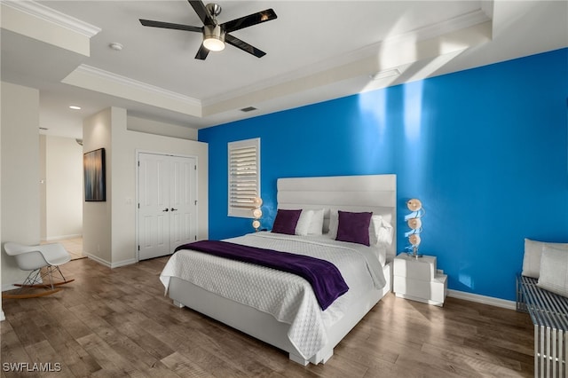 bedroom with hardwood / wood-style floors, a raised ceiling, ceiling fan, ornamental molding, and a closet