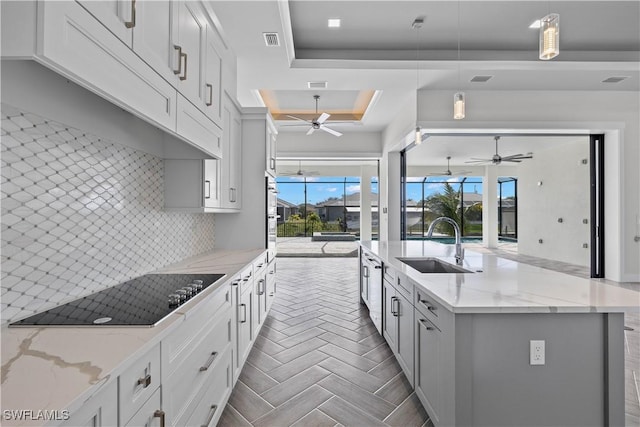 kitchen featuring light stone countertops, a raised ceiling, an island with sink, pendant lighting, and black electric cooktop