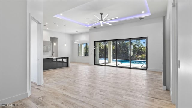 unfurnished living room featuring a raised ceiling, ceiling fan, a high ceiling, and light wood-type flooring