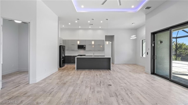 kitchen with gray cabinets, pendant lighting, an island with sink, a raised ceiling, and black fridge