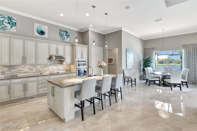 kitchen featuring light stone counters, double oven, decorative light fixtures, a breakfast bar area, and a center island with sink